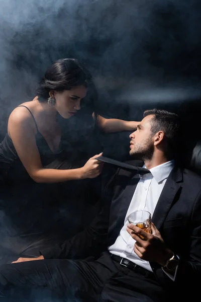 Bruna donna tirando cravatta di fidanzato con whisky su sfondo nero con fumo — Foto stock