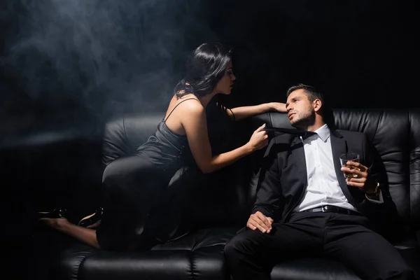 Man holding glass of whiskey near passionate woman in dress on couch on black background with smoke — Stock Photo