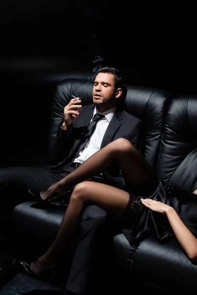 Чоловік у костюмі тримає цигарку біля чуттєвої жінки на дивані ізольовано на чорному — стокове фото