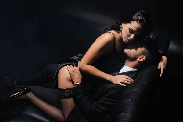 Чоловік у костюмі торкається чуттєвої дівчини брюнетки на дивані на чорному тлі — стокове фото
