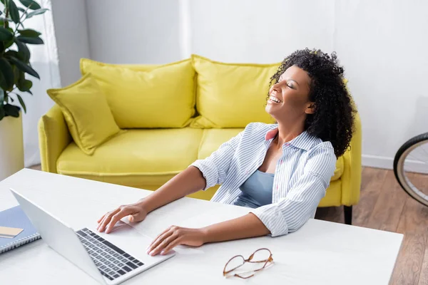 Счастливый африканский американский блогер сидит рядом с ноутбуком, очки и кредитная карта на столе — стоковое фото