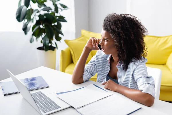 Freelancer afro-americano confuso falando em smartphones perto de notebooks e laptop — Fotografia de Stock