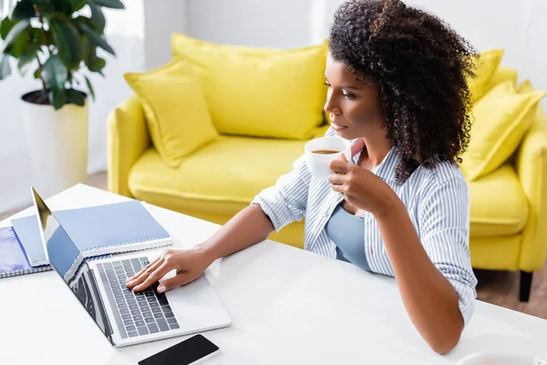 Telelavoro afroamericano con tazza di caffè utilizzando laptop con schermo bianco a casa — Foto stock