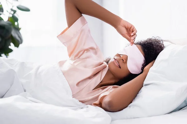 African american woman in silk pajama holding sleep mask on bed — Stock Photo