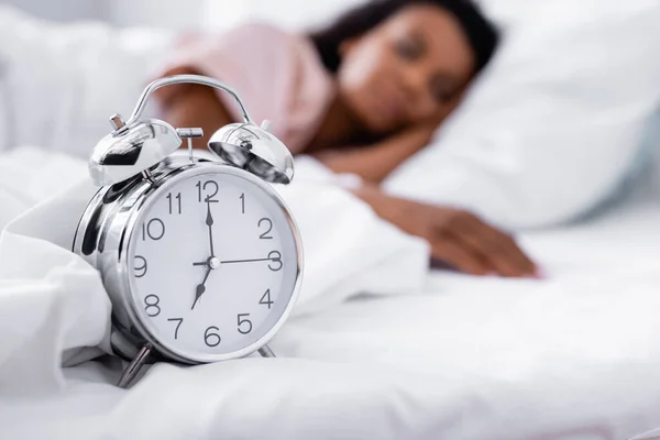 Alarm clock on white bedding near african american woman on blurred background — Stock Photo
