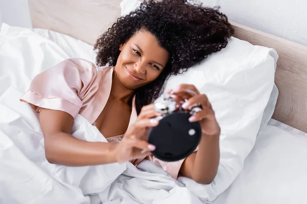 Mulher americana africana alegre segurando despertador em primeiro plano desfocado na cama — Fotografia de Stock