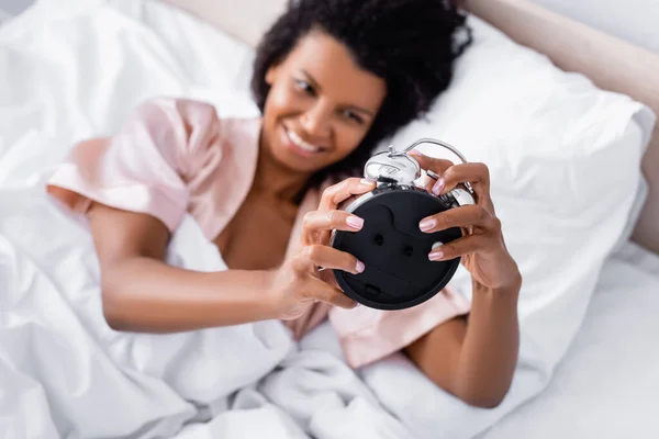 Alarm clock in hands of african american woman on blurred background on bed — Stock Photo