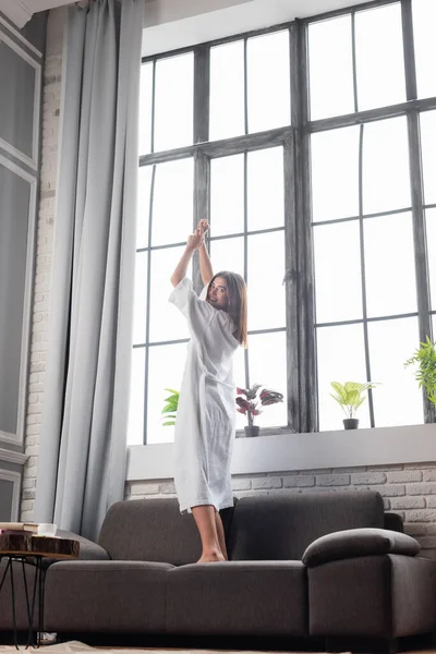 Smiling young adult woman in bathrobe standing on couch with hands in air at home — Stock Photo