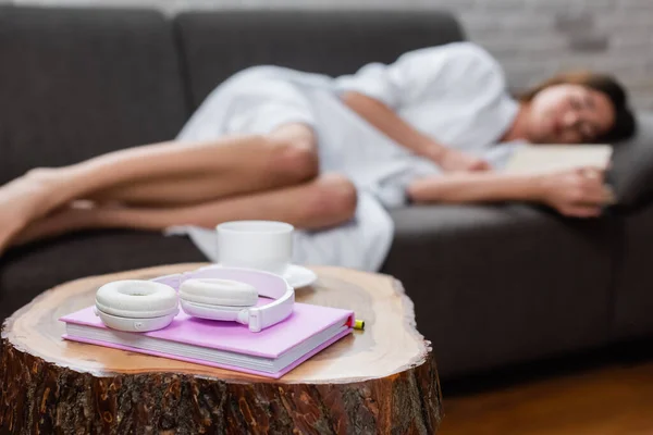 Headphones, cup of coffee and notebook on blurred background with sleeping young adult woman at home — Stock Photo