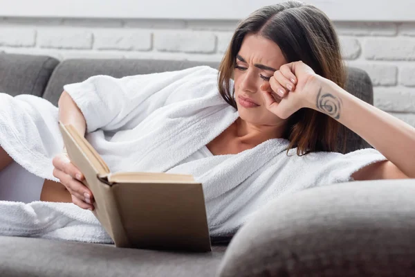 Mujer adulta joven molesta leyendo libro en el sofá gris en casa - foto de stock