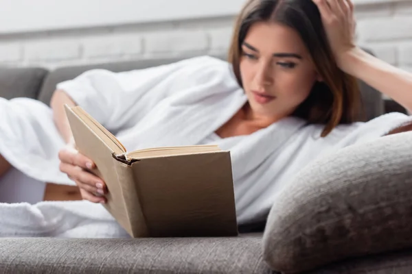 Mujer adulta joven en el libro de lectura de albornoz en el sofá gris en casa - foto de stock