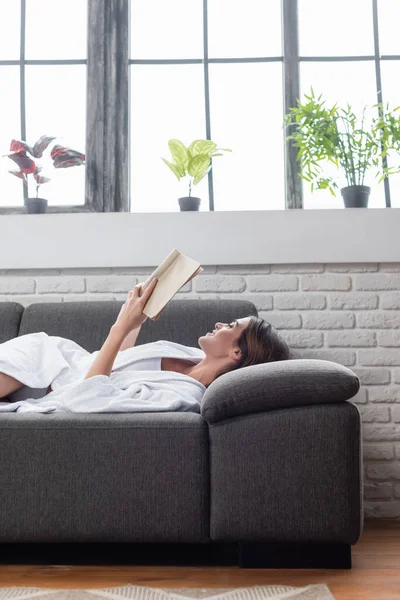 Jeune femme adulte souriante en peignoir blanc livre de lecture sur canapé gris à la maison — Photo de stock