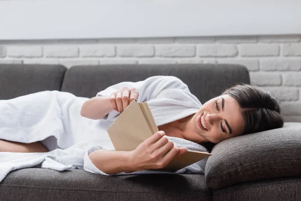 Happy young adult woman in white bathrobe reading book on grey couch at home — Stock Photo