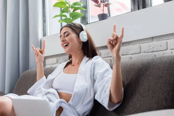 Молодая взрослая женщина с рок-жестом сидит со скрещенными ногами и слушает музыку в наушниках в гостиной — стоковое фото