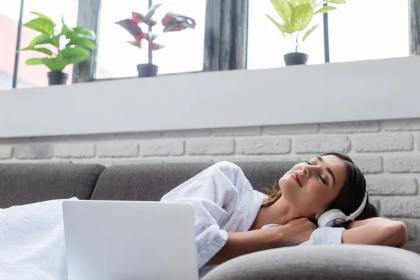 Peaceful young adult woman with closed eyes in headphones resting on couch near laptop at home — Stock Photo