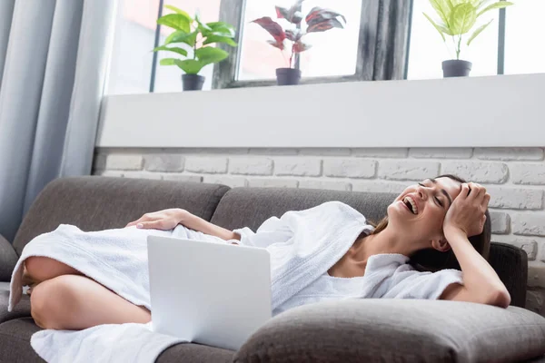 Laughing young adult woman with closed eyes resting on couch near laptop at home — Stock Photo