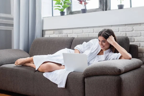 Sorrindo jovem mulher adulta deitada no sofá com laptop em casa — Fotografia de Stock