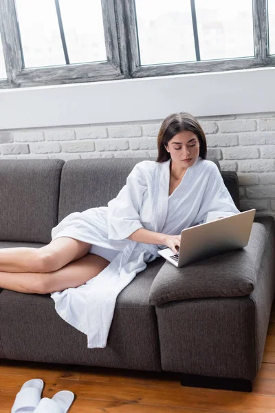 Serious young adult woman in bathrobe lying on couch and using laptop at home — Stock Photo