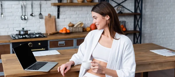 Happy young adult woman in white bathrobe sitting near laptop in loft, banner — Stock Photo