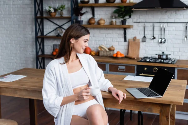 Smiling young adult woman in white bathrobe sitting near laptop in loft — Stock Photo