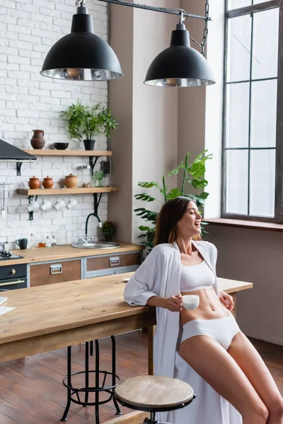 Positive jeune femme adulte en peignoir blanc tenant tasse de café dans le loft moderne — Photo de stock