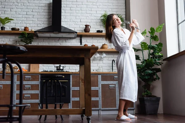 Playful young adult woman in white bathrobe holding spatula and singing in modern loft — Stock Photo