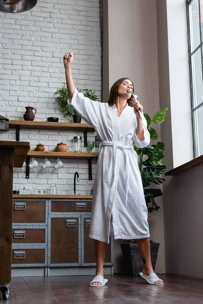 Playful young adult woman in white bathrobe holding spatula and singing with hand in air gesture in modern loft — Stock Photo