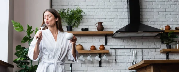 Jeune femme adulte ludique en peignoir blanc tenant tasse de café et spatule et chantant dans le loft moderne, bannière — Photo de stock