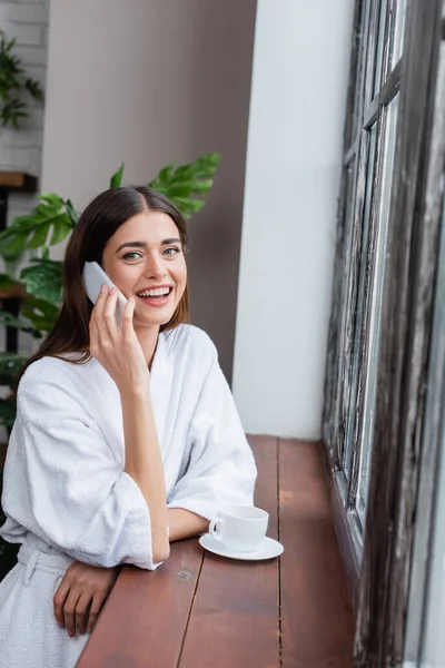 Smiling young adult woman speaking on cellphone near window in living room — Stock Photo