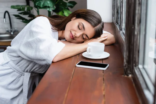 Mujer adulta joven durmiendo cerca del teléfono celular y taza de café en el alféizar en la sala de estar - foto de stock