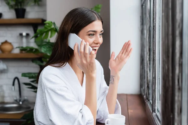 Sonriente mujer adulta joven hablando por teléfono celular y haciendo un gesto con la mano cerca de la ventana en la sala de estar - foto de stock