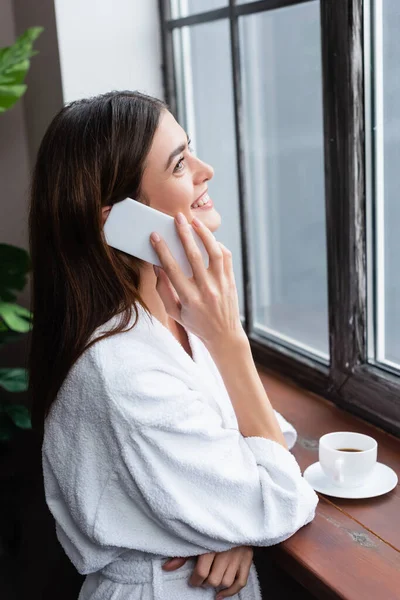 Sonriente mujer joven adulta en albornoz hablando por teléfono celular y mirando por la ventana en la sala de estar - foto de stock