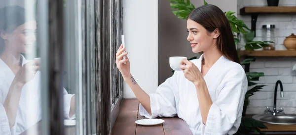 Усміхнена молода доросла жінка в халаті бере селфі на мобільний телефон біля вікна в сучасному лофт, банер — стокове фото
