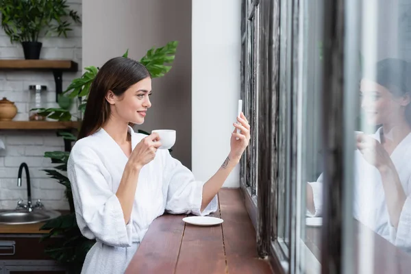 Hübsche junge erwachsene Frau im Bademantel macht Selfie mit Handy am Fenster in modernem Dachboden — Stockfoto