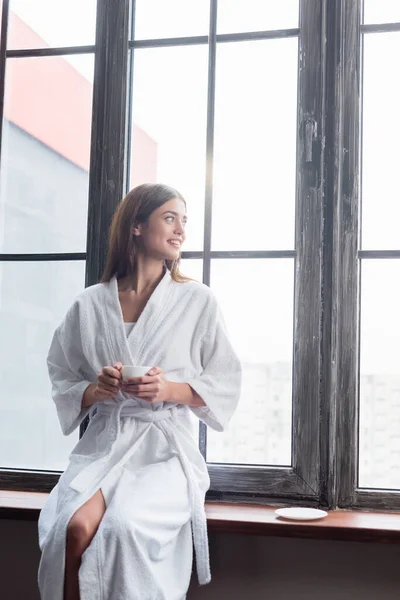 Pretty young adult woman in bathrobe sitting with cup of coffee near window at home — Stock Photo