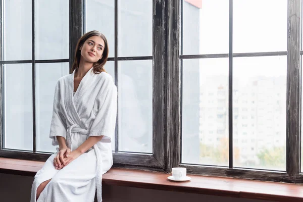 Pretty young adult woman in bathrobe sitting on sill with cup of coffee near window at home — Stock Photo