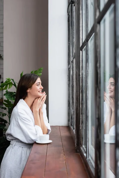 Sorridente jovem mulher adulta olhando para fora janela com as mãos perto do rosto na sala de estar moderna — Fotografia de Stock