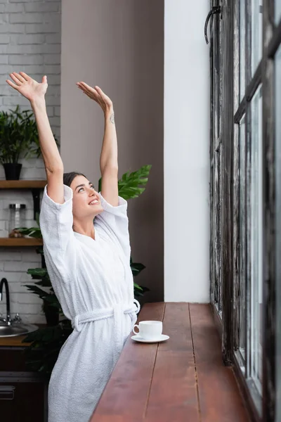 Happy young adult woman in bathrobe standing near window with hands in air in modern living room — Stock Photo