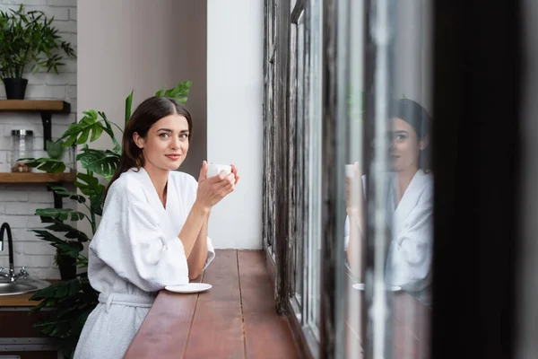 Feliz jovem mulher adulta segurando xícara de café perto da janela com reflexão na sala de estar moderna — Fotografia de Stock