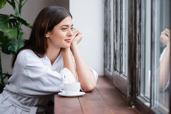 Sonriente joven mujer adulta sentada con las manos cerca de la cara y mirando por la ventana en casa - foto de stock