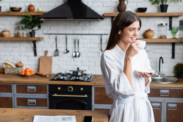 Smiling young adult woman in bathrobe drinking coffee near table with newspaper and cellphone in modern loft — Stock Photo
