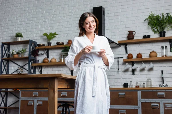 Glückliche junge erwachsene Frau im Bademantel, die Kaffee trinkt und im modernen Loft in die Kamera blickt — Stockfoto