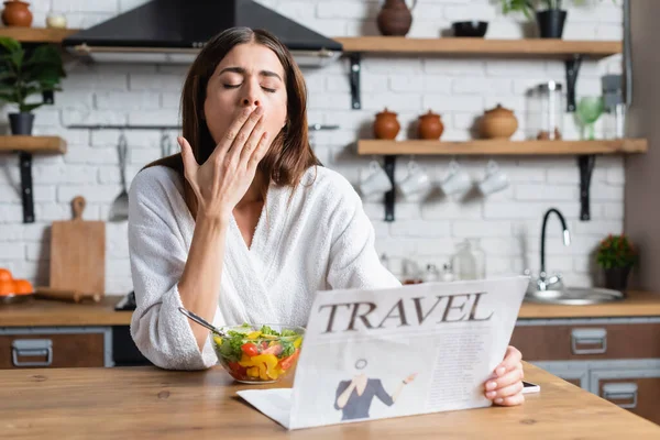 Jovem mulher adulta sonolenta em roupão comendo salada e lendo jornal de viagem na cozinha moderna — Fotografia de Stock
