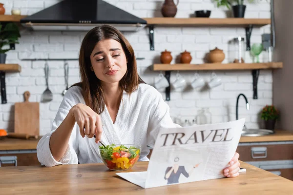 Mujer joven y aburrida en albornoz comiendo ensalada y leyendo el periódico de viaje en la cocina moderna - foto de stock