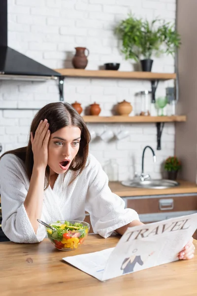 Eccitato giovane donna adulta in accappatoio mangiare insalata e leggere giornale di viaggio in cucina moderna — Foto stock
