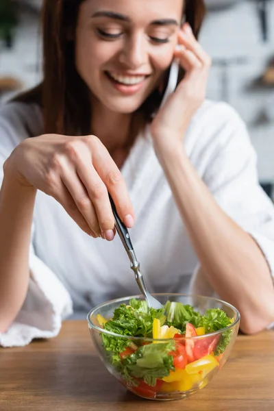 Souriant jeune femme adulte en peignoir manger de la salade et parler sur téléphone portable dans la cuisine — Photo de stock