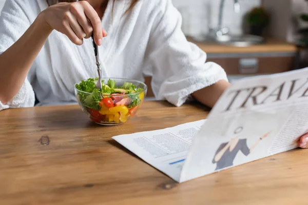 Partial view of young adult woman in bathrobe eating salad and reading travel newspaper in kitchen — Stock Photo