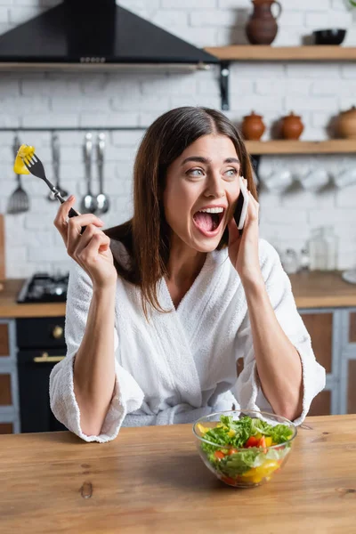 Young adult woman in bathrobe eating vegetables salad and speaking on cellphone in modern kitchen — Stock Photo