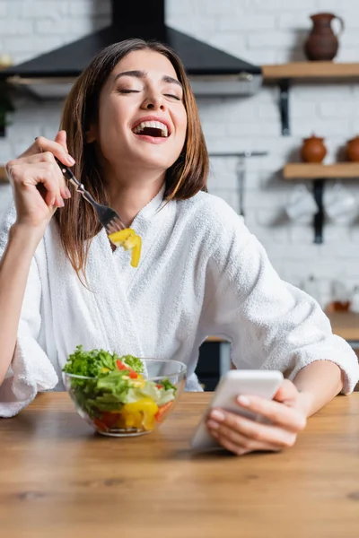 Rire jeune femme adulte en peignoir manger salade de légumes et en utilisant un téléphone portable dans la cuisine moderne — Photo de stock