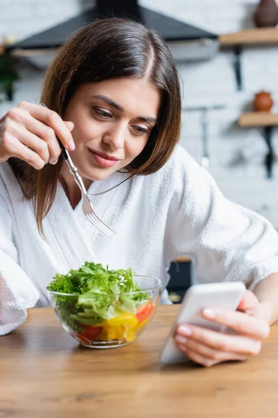 Sorrindo jovem mulher adulta em roupão comendo salada de legumes e olhando para o celular na cozinha moderna — Fotografia de Stock